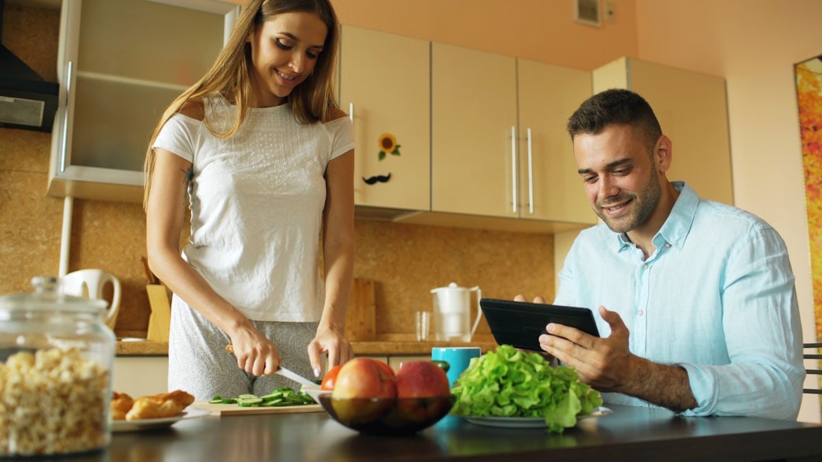 Attractive couple chatting in the kitchen early morning. Handsome man using tablet while his girlfriend cooking breakfast at home